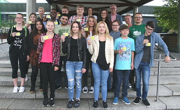Große Gruppe junger Menschen steht auf einer Treppe vor einem Gebäude und blickt in die Kamera. Einige heben gelbe Ausweise oder Karten hoch. Wirkt wie ein Gruppenfoto einer Klasse oder eines Kurses.