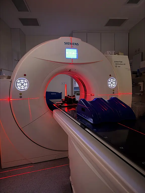 Großer Computertomograph in einem Krankenhausraum: runde Öffnung mit leuchtend roten Laserlinien zur Positionierung, davor ein verschiebbarer Patiententisch mit Halterung. Das Gerät ist eingeschaltet und bereit für eine CT‑Untersuchung.