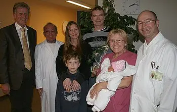 Eine Gruppe von sieben Personen steht in einem Raum. In der Mitte hält eine Frau ein neugeborenes Baby. Um sie herum stehen ein Junge, ein Mann und mehrere Ärzte in weißen Kitteln. Alle wirken glücklich und feiern einen besonderen Moment.