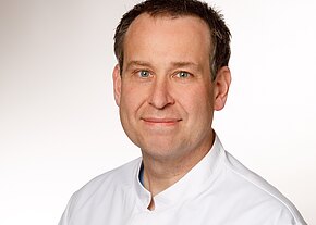 Dr. med. Stefan Pielmeier Porträt eines Mannes mittleren Alters mit kurzem braunem Haar. Er schaut lächelnd in die Kamera und trägt einen weißen Kittel, vor neutral hellem Hintergrund.