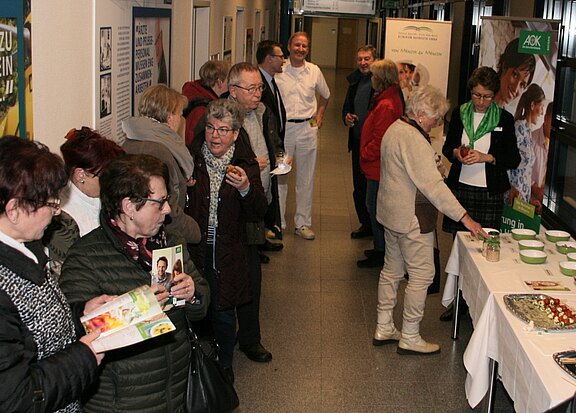 Menschen versammeln sich in einem Flur bei einer Gesundheits-Infoveranstaltung; sie stehen an einem Tisch mit Schalen und Speisen an, blättern in Broschüren und sprechen miteinander. Eine Betreuerin hilft am Stand; im Hintergrund weitere Besucher.