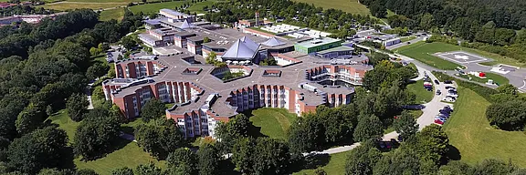 Luftaufnahme eines modernen Gebäudekomplexes, umgeben von Bäumen und Wiesen. Der Gebäudekomplex hat eine ungewöhnliche, sternförmige Architektur und beherbergt mehrere Flügel. Im Hintergrund sind weitere Einrichtungen und ein Landeplatz für Hubschrauber zu sehen.