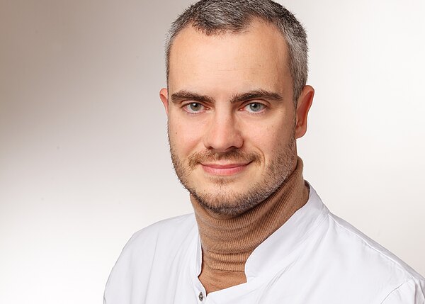 Porträt eines jungen Mannes mit kurz geschnittenem grauem Haar und Bartstoppeln. Er trägt einen weißen Kittel über einem braunen Rollkragen und lächelt leicht in die Kamera. Neutraler, heller Hintergrund.