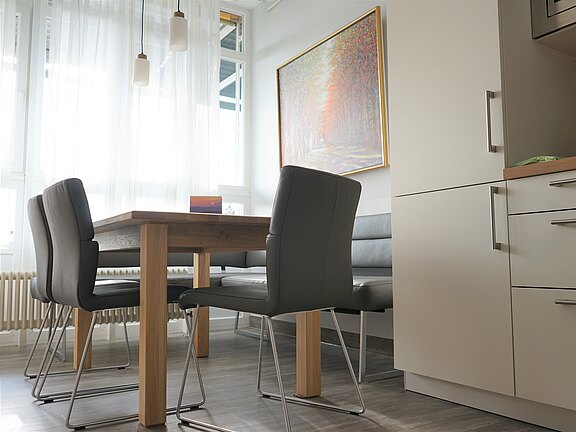 Heller Essbereich in moderner Küche: Holztisch mit grauen, gepolsterten Stühlen und Eckbank, daneben helle Einbauküchenfront. Große Fenster mit transparenten Vorhängen lassen viel Licht herein; über dem Tisch hängen kleine Pendelleuchten, an der Wand ein farbiges Bild.