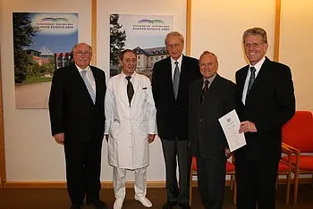 Fünf Personen stehen in einem Innenraum, darunter ein Arzt in einem weißen Kittel. Sie lächeln und scheinen an einem feierlichen Anlass teilzunehmen. Im Hintergrund sind Plakate sichtbar, die eine Einrichtung zeigen.