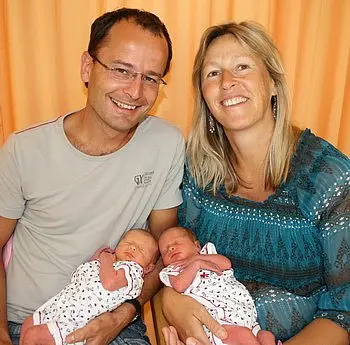 Eine glückliche Familie sitzt zusammen und lächelt. Ein Vater und eine Mutter halten jeweils ein Neugeborenes in ihren Armen. Die Eltern tragen lässige Kleidung und strahlen Freude aus. Im Hintergrund ist ein orangener Vorhang zu sehen.