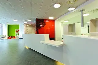 Eine moderne Empfangshalle mit einem weißen Empfangstresen. Im Hintergrund sind Wände in lebhaften Farben, darunter ein rot lackierter Akzent. Zu sehen sind anthrazitfarbene Fliesen und eine gemütliche Sitzgelegenheit in einer Ecke. Die Atmosphäre ist hell und einladend.