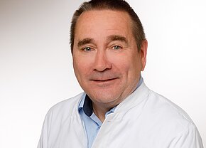 Dr. med. René Tandler Ein mittelalterlicher Mann mit einem freundlichen Lächeln trägt einen weißen Kittel über einem blauen Hemd. Er schaut in die Kamera und hat kurzes, leicht graues Haar. Der Hintergrund ist hell und unauffällig.