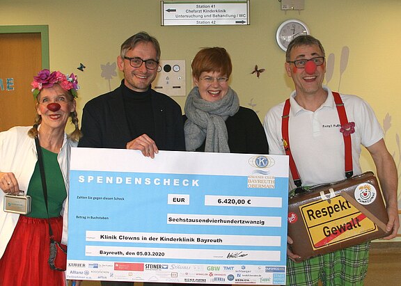 Vier Erwachsene, darunter zwei als Clowns, halten einen großen Spendencheck. Der Betrag: 6.420 Euro für die Klinik-Clowns in der Kinderklinik Bayreuth. Fröhliche Stimmung bei der Spendenübergabe.