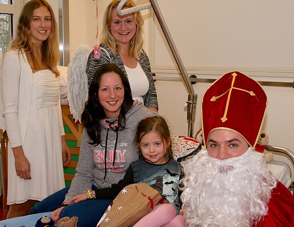 Ein kleines Mädchen sitzt lächelnd zwischen seiner Mutter und einem als Nikolaus verkleideten Besucher mit roter Mitra und weißem Bart. Zwei als Engel verkleidete junge Frauen stehen dahinter. Fröhliche, weihnachtliche Besuchsszene.