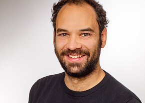 Stefan Knauer Porträt eines lächelnden erwachsenen Mannes mit dunklem, leicht lockigem Haar und Bart. Er trägt ein dunkles Shirt und schaut direkt in die Kamera, vor neutralem Hintergrund.