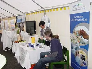 Das Bild zeigt einen Informationsstand in einem Zelt. Eine Person steht am Tisch und erklärt etwas, während eine andere Person sitzt und zuhört. Auf dem Tisch sind verschiedene Informationsmaterialien und Utensilien ausgebreitet. Im Hintergrund sind Banner mit Informationen über das Unternehmen zu sehen.