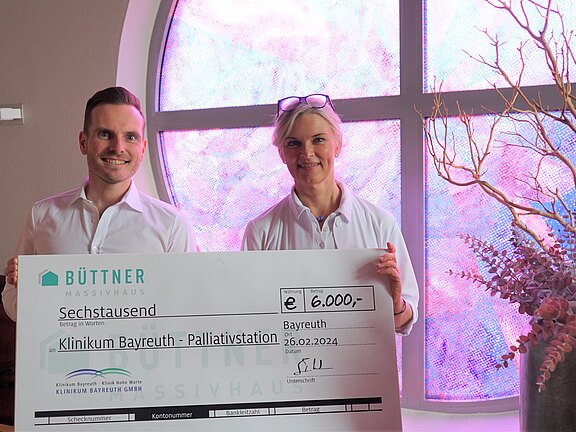 Zwei Personen präsentieren einen großen Spendenscheck: 6.000 Euro für die Palliativstation des Klinikums Bayreuth, datiert 26.02.2024. Der Scheck stammt von Büttner Massivhaus.