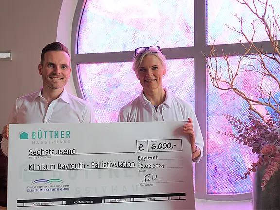 Zwei Personen präsentieren einen großen Spendenscheck: 6.000 Euro für die Palliativstation des Klinikums Bayreuth, datiert 26.02.2024. Der Scheck stammt von Büttner Massivhaus.