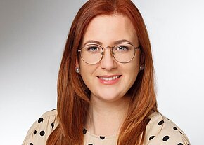 Porträtfoto einer lächelnden Frau mit langen roten Haaren und runden Brillen. Sie trägt eine helle Bluse mit schwarzen Punkten. Neutrale, helle Hintergrundfläche; Fokus auf ihr freundliches Gesicht.