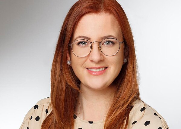 Porträtfoto einer lächelnden Frau mit langen roten Haaren und runden Brillen. Sie trägt eine helle Bluse mit schwarzen Punkten. Neutrale, helle Hintergrundfläche; Fokus auf ihr freundliches Gesicht.
