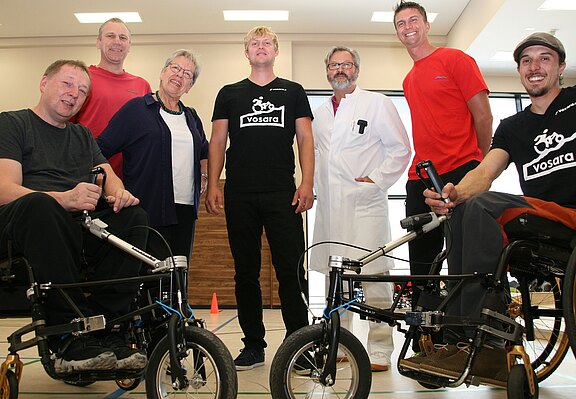 Gruppenfoto in einem Innenraum: Zwei Personen im Rollstuhl mit angekoppelten Handbike-Vorderrädern; daneben mehrere stehende Unterstützer, darunter ein Mann im weißen Kittel. Alle lächeln. Die Szene wirkt wie eine Präsentation oder ein Teamfoto zu barrierefreier Mobilität.