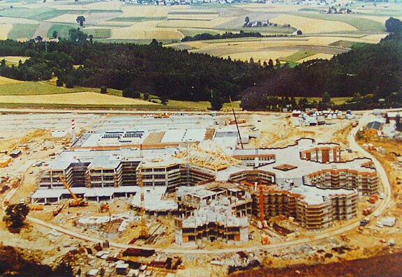 Luftaufnahme einer großen Baustelle auf dem Land: Mehrflügeliger Gebäudekomplex im Rohbau mit mehreren Stockwerken, Kränen und Erdhügeln. Straßen und Zufahrten umgeben die Anlage. Im Hintergrund Felder und Wald.