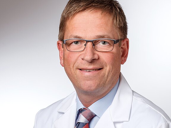 Porträt eines mittelalten Mannes mit Brille in weißem Laborkittel und Krawatte, freundlich lächelnd vor neutralem Hintergrund.