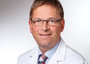 Porträt eines mittelalten Mannes mit Brille in weißem Laborkittel und Krawatte, freundlich lächelnd vor neutralem Hintergrund.