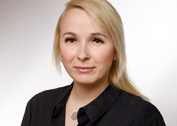 Porträt einer Person mit langem blondem Haar vor hellem Hintergrund. Sie trägt ein schwarzes Hemd und eine silberne Kette. Neutrale Mimik, Blick in die Kamera. Kopf- und Schulteraufnahme.