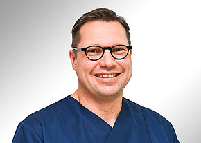 Porträt eines lächelnden Mannes mit runder schwarzer Brille und kurz geschnittenen Haaren. Er trägt dunkelblaue medizinische Kleidung und blickt direkt in die Kamera vor hellem, neutralem Hintergrund.