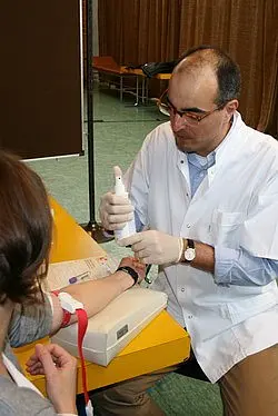 Ein Arzt führt einen Bluttest an einer Patientin durch. Er hält ein Instrument in der Hand, während die Patientin am Arm eine Blutabnahme erhält. Beide Personen sind konzentriert auf den Vorgang.