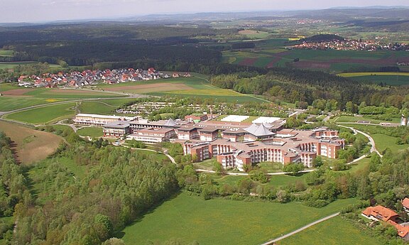 Luftaufnahme eines großen Gebäudekomplexes mit mehreren Flügeln, eingebettet in Wiesen, Felder und Wald. Im Hintergrund liegen verstreute Ortschaften und sanfte Hügel. Der Fokus liegt auf der weitläufigen Anlage inmitten der grünen Landschaft.