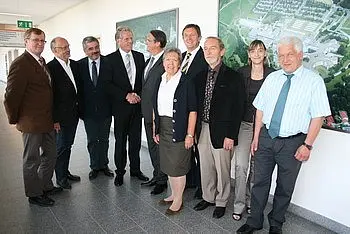 Eine Gruppe von elf Personen steht in einem Flur. Sie lächeln und posieren für ein Foto. Einige tragen Anzüge, andere formelle Kleidung. Im Hintergrund ist eine Wand mit Bildern zu sehen. Die Atmosphäre wirkt freundlich und einladend.