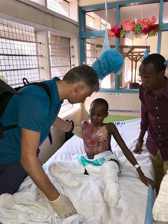 Im Krankenhausbett sitzt ein verletztes Kind mit großflächigen Verbrennungen am Oberkörper und bandagierten Beinen. Eine behandschuhte medizinische Fachkraft untersucht es behutsam, eine weitere Person steht unterstützend daneben.