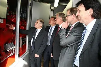 Eine Gruppe von fünf Männern in Anzügen steht vor einem technischen Gerät. Ein Mann zeigt auf die Maschine, während die anderen aufmerksam beobachten. Die Szene vermittelt Interesse und Diskussion über die Technologie im Hintergrund.