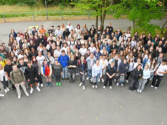 Eine große Gruppe von Menschen steht im Freien, auf einem gepflasterten Platz. Die Personen sind vielfältig gekleidet und lächeln in die Kamera. Bäume und Wiese sind im Hintergrund sichtbar. Die Anordnung der Gruppe vermittelt ein Gefühl von Gemeinschaft und Zusammengehörigkeit.