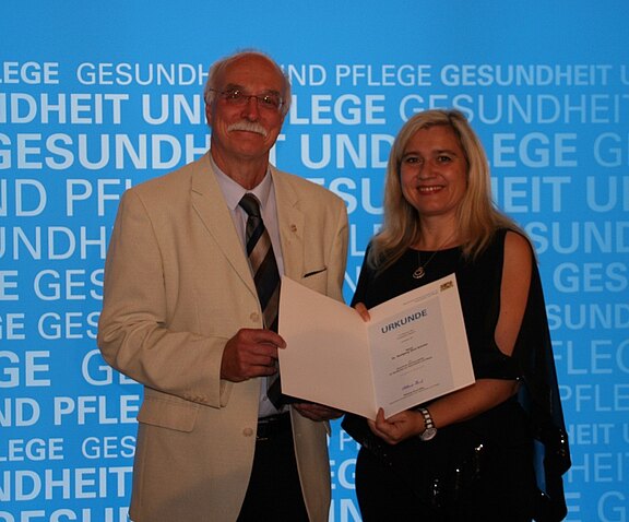 Ein Mann im hellen Anzug und eine Frau im schwarzen Kleid stehen nebeneinander und halten gemeinsam eine Urkunde. Beide lächeln. Hintergrund zeigt wiederholt die Worte „Gesundheit und Pflege“. Szene wirkt wie eine Auszeichnung oder Preisverleihung.