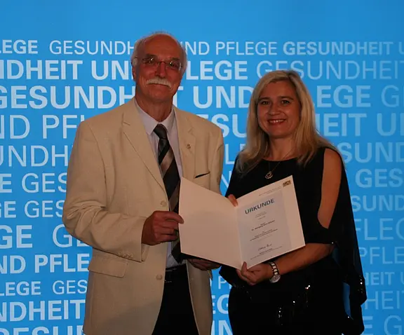 Ein Mann im hellen Anzug und eine Frau im schwarzen Kleid stehen nebeneinander und halten gemeinsam eine Urkunde. Beide lächeln. Hintergrund zeigt wiederholt die Worte „Gesundheit und Pflege“. Szene wirkt wie eine Auszeichnung oder Preisverleihung.