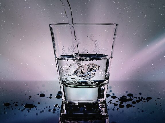 Klares Trinkglas, in das Wasser gegossen wird. Ein Diamant im Glas verursacht Spritzer; Tropfen liegen auf der spiegelnden Oberfläche.