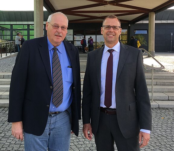 Zwei Männer in formeller Kleidung stehen nebeneinander vor einem Gebäudeeingang mit Treppe und Vordach. Sie blicken in die Kamera und lächeln freundlich.