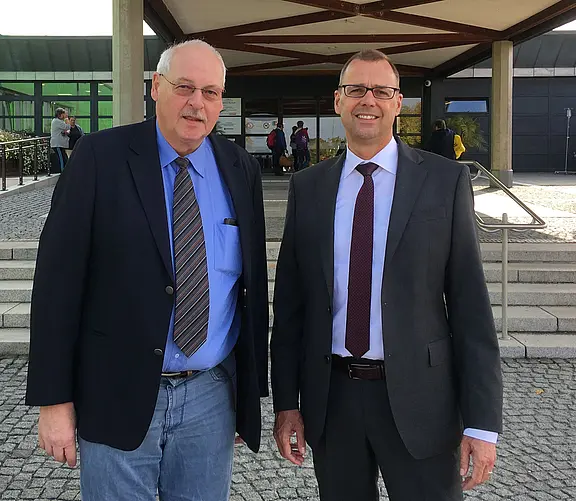Zwei Männer in formeller Kleidung stehen nebeneinander vor einem Gebäudeeingang mit Treppe und Vordach. Sie blicken in die Kamera und lächeln freundlich.