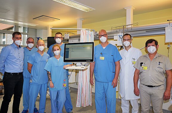Neun Krankenhausmitarbeitende in OP-Kleidung, Kitteln und Masken stehen in einem Behandlungsraum. In der Mitte befindet sich ein Monitor auf einem Wagen mit geöffneter Software. Gruppenfoto, das Team zeigt seine medizinische Technik.