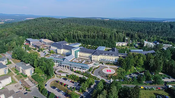 Luftaufnahme eines großen Krankenhauskomplexes inmitten von Wald. Mehrere Gebäude sind durch Wege verbunden, vorne Parkplätze und eine Zufahrtsstraße. Zentral liegt ein markierter Hubschrauberlandeplatz mit rotem Kreuz. Im Hintergrund bewaldete Hügel unter klarem Himmel.