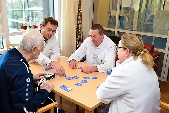 Vier Personen sitzen an einem Tisch und spielen mit blauen Karten. Eine ältere Person, die ein medizinisches Gerät in der Hand hält, wird von drei Fachkräften in weißen Kitteln betreut. Die Atmosphäre wirkt freundlich und kommunikativ, während sie gemeinsam an der Aktivität arbeiten.