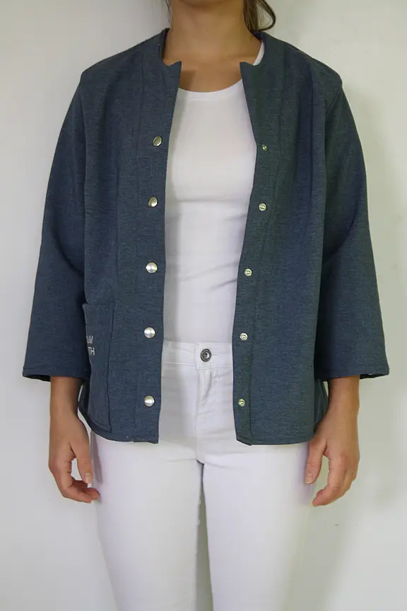 Person trägt offene, dunkelgraublaue Jacke mit Druckknöpfen und 3/4-Ärmeln über weißem T‑Shirt und weißer Hose. Fokus auf der Jacke, vorne offen, schlichte, gerade Form.
