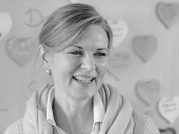 Schwarzweiß-Porträt einer lächelnden Frau, leicht seitlich zum Betrachter gewandt. Sie trägt Hemd und Strickjacke, die Haare sind zusammengebunden. Im Hintergrund unscharfe herzförmige Dekoration; der Fokus liegt auf ihrer freundlichen Mimik.