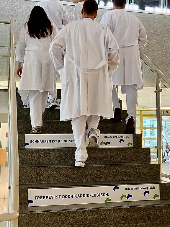Mehrere Personen in weißen Kitteln steigen eine Treppe hinauf. Auf den Stufen stehen motivierende Sprüche wie „Treppe? Ist doch kardio-logisch“ und „Schnaufen ist keine Schande“. Die Szene wirbt für Bewegung und die gesundheitlichen Vorteile des Treppensteigens.