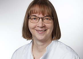 Porträt einer lächelnden Frau mit schulterlangem braunen Haar und Brille, in weißem medizinischen Kittel, vor hellem neutralem Hintergrund.