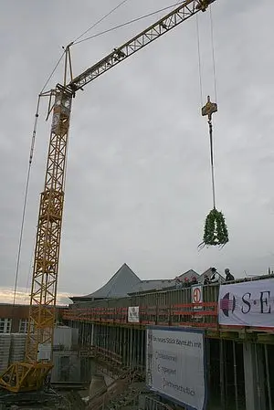 Ein Kran hebt einen Kranz über eine Baustelle, während Arbeiter auf dem Gerüst stehen. Im Hintergrund sind Teile eines Gebäudes und Werbebanner zu sehen. Der Himmel ist bewölkt und sorgt für eine düstere Atmosphäre.