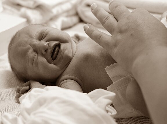 Ein weinendes Baby liegt auf einer weichen Unterlage. Eine Hand berührt sanft das Baby, das Unbehagen zeigt. Die Szene vermittelt eine emotionale Verbindung zwischen dem Baby und der beruhigenden Hand.