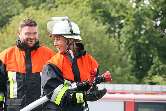 Zwei Feuerwehrleute in Schutzkleidung im Freien. Eine Feuerwehrfrau mit Helm hält einen Wasserschlauch mit Sprühdüse und lächelt, während ein Kollege daneben steht. Eindruck von Übung oder Einsatzvorbereitung; im Hintergrund angedeutet ein Feuerwehrfahrzeug, umgeben von Bäumen.