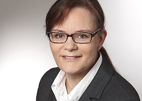 Professionelles Porträt: Frau mit Brille, braunen Haaren und Pferdeschwanz, in dunklem Blazer und weißer Bluse, lächelt leicht vor neutralem Hintergrund.