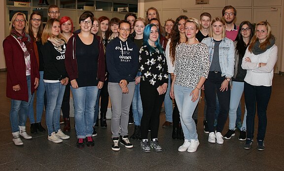 Gruppenfoto junger Erwachsener in einem Innenraum. Etwa zwanzig Personen stehen dicht beisammen und schauen zur Kamera, überwiegend lässig gekleidet. Stimmung freundlich, einige lächeln. Neutraler Hintergrund mit Türen. Schwerpunkt: Gemeinschaftsbild einer Klasse oder Gruppe.