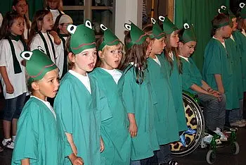 Eine Gruppe von Kindern steht in einer Reihe und trägt grüne Gewänder sowie Froschkappen. Sie wirken begeistert und singen zusammen. Im Hintergrund sind weitere Kinder und eine Person im Rollstuhl zu sehen.